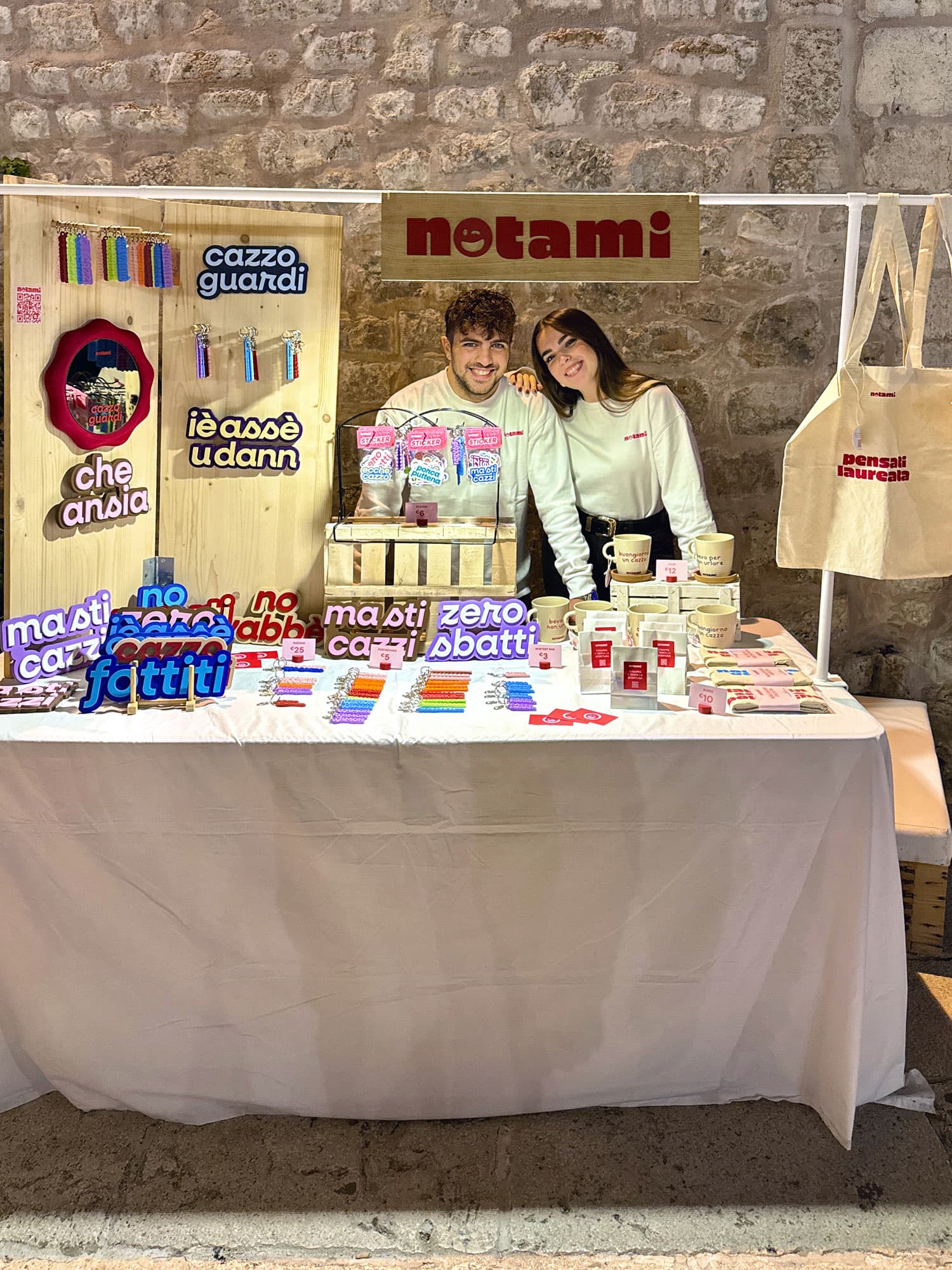 Vanni e Samantha sorridenti dietro al banco Notami a un mercatino, tra insegne colorate stampate in 3D, portachiavi, tazze e una shopper