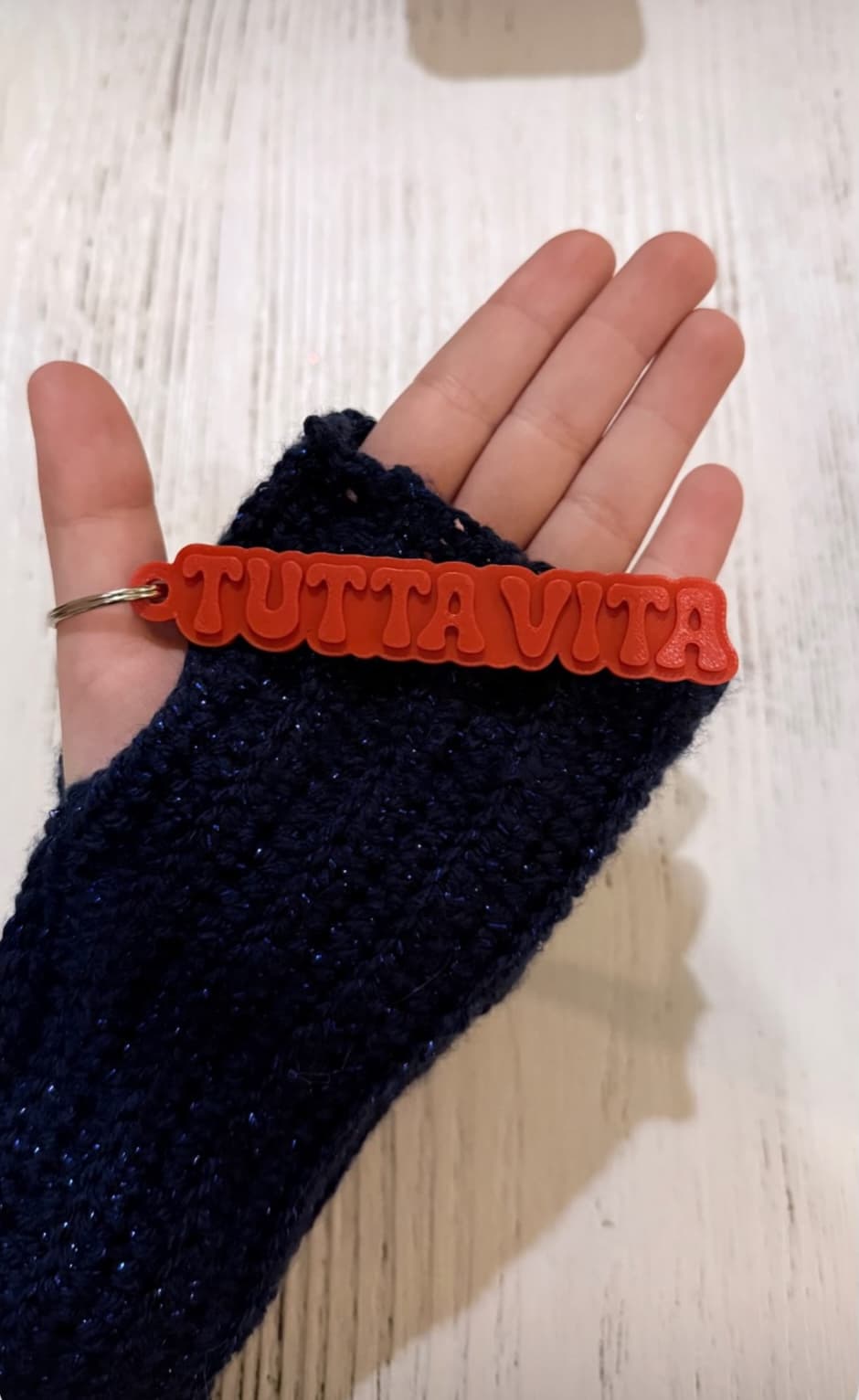 Hand in a blue knitted glove holding the red keychain reading “TUTTAVITA”