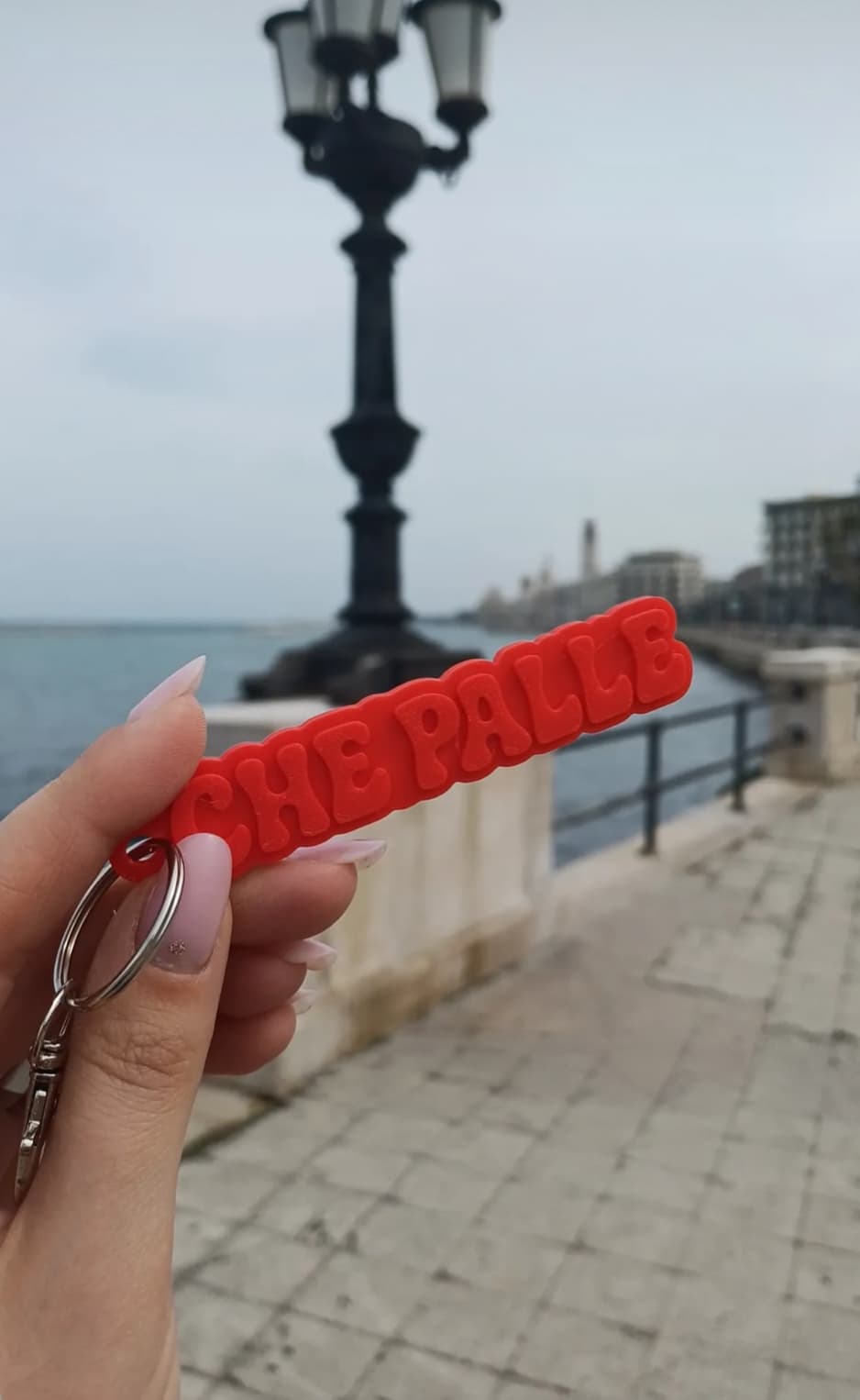 Red keychain reading “CHE PALLE” held up on the seafront